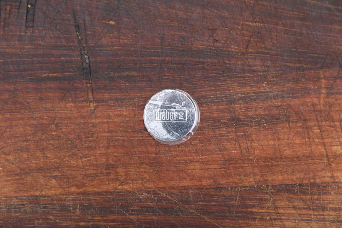 A small metal disc with the Weber logo on, sat on a wooden table.