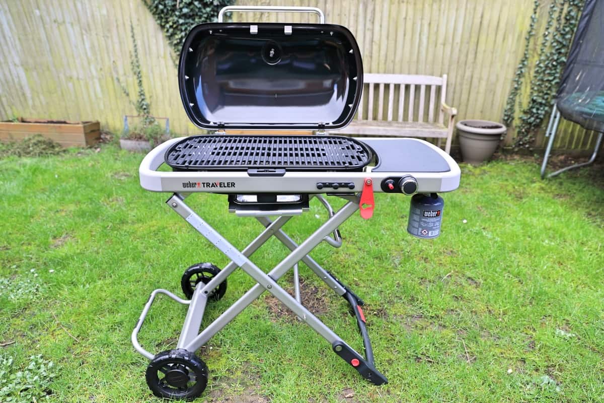 Weber Traveler with lid open, on a grass lawn, in front of a wooden fe.