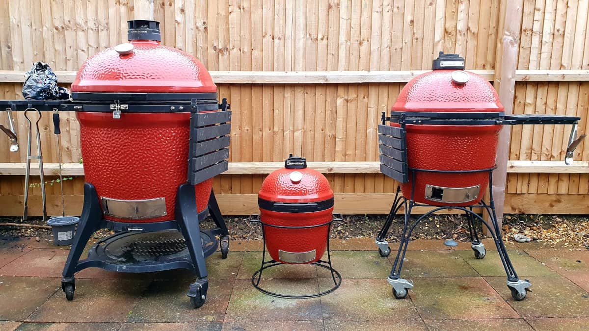 All three sizes of Kamado Joe smokers side by side in front of a wooden paneled fence.