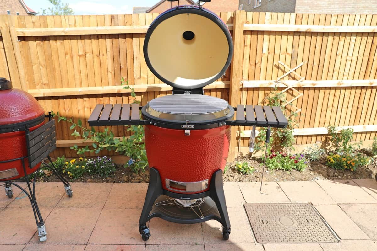 kamado joe big joe III bathed in sunlight, in front of a wooden fence, on a paved pa.