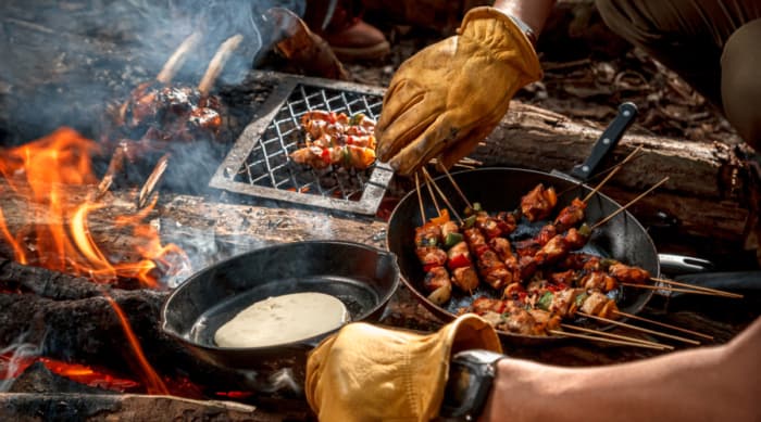 A man with gloves outdoor cooking, using cast iron pans and various BBQ tools.