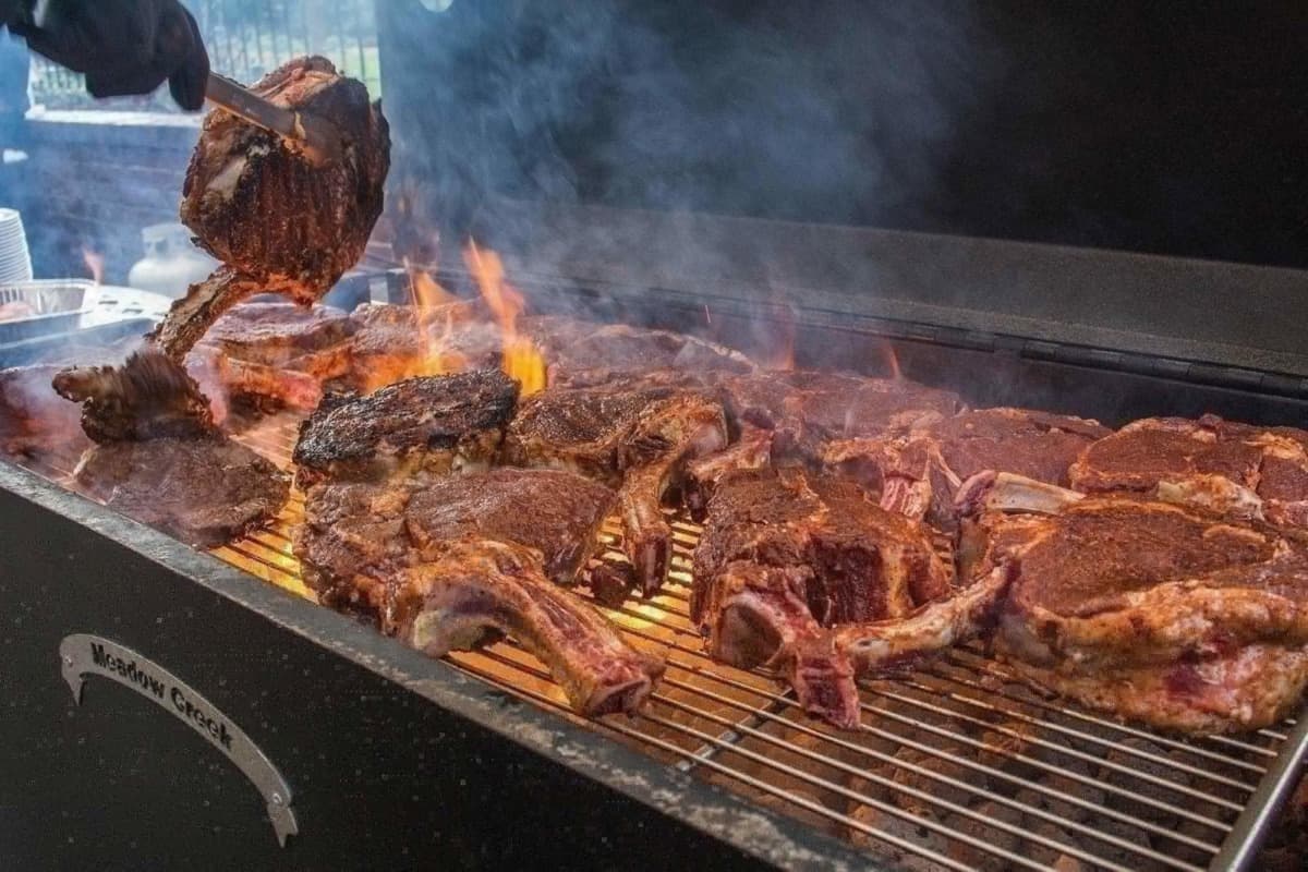 A Meadow Creek BBQ26 being used to cook numerous tomahawk steaks, with flames leaping up through the grate