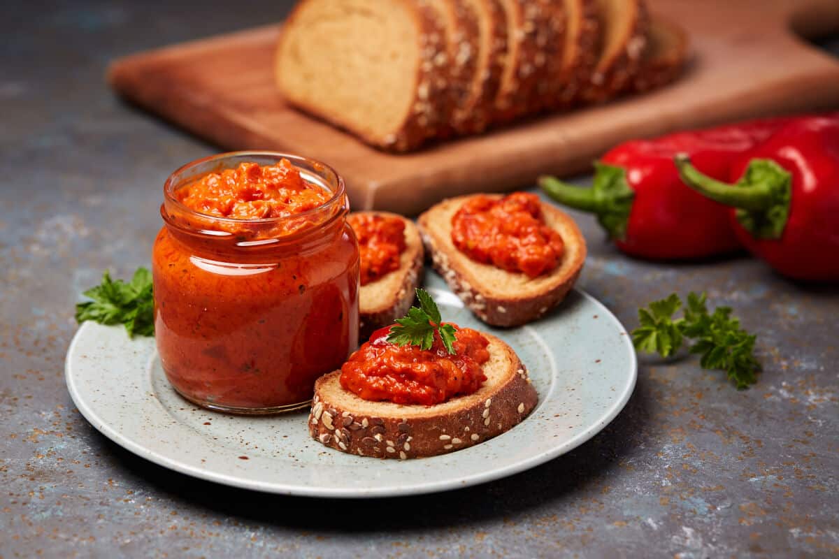 A jar of ajvar on a white plate, next to some slices of seeded bread with ajvar spread on top and a parsley leaf on top of t.