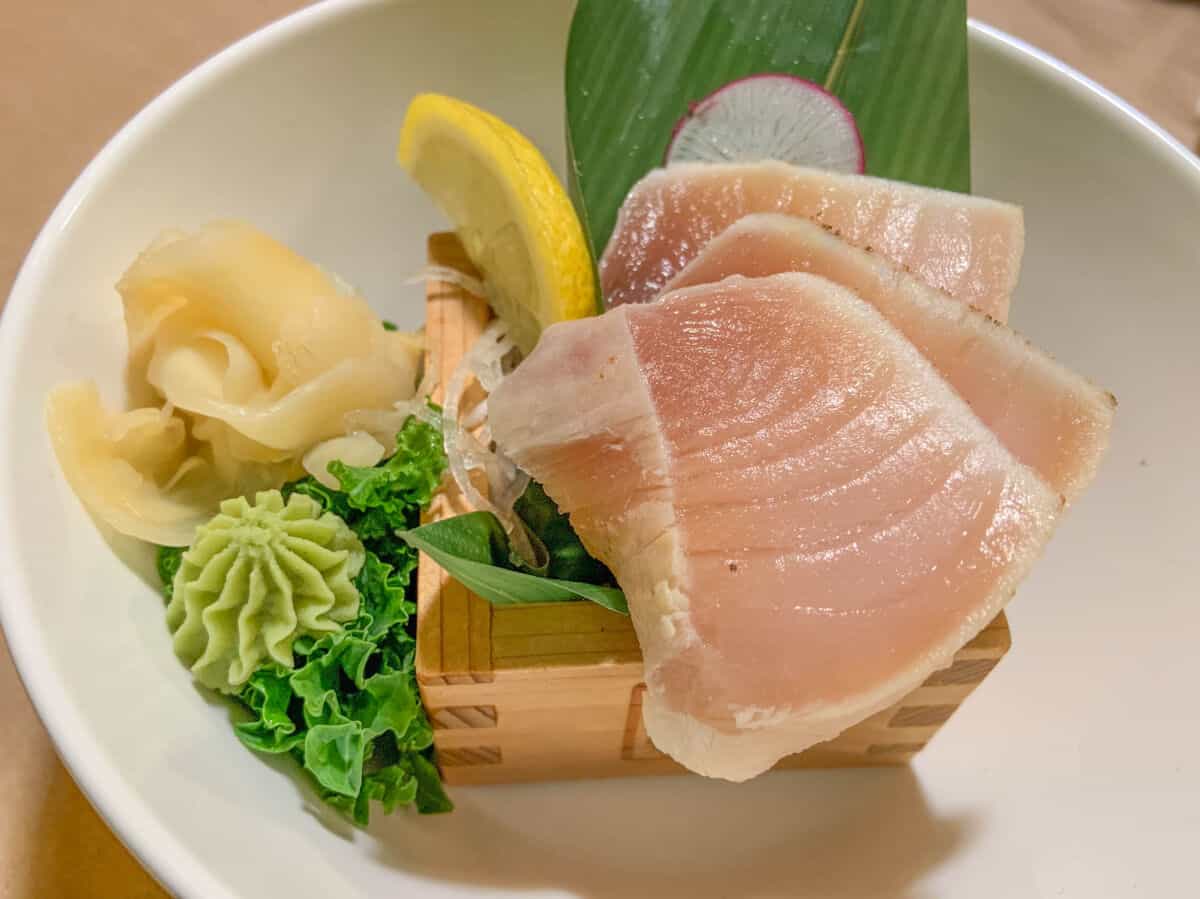 Slices of albacore tuna in a white bowl, with some radish, dumplings, lemon and gre.