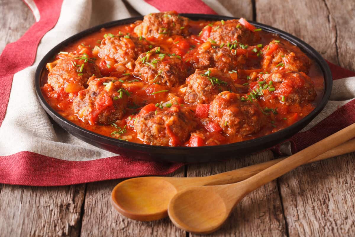 A black plate full of albondigas meatballs, on a red and white tea towel, with two wooden spoons in fr.