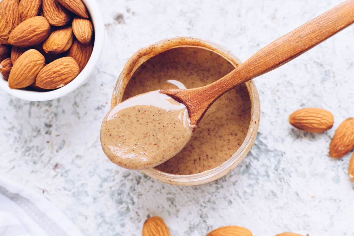 A bowl of almond butter next to a bowl of whole almonds on a light colored ta.