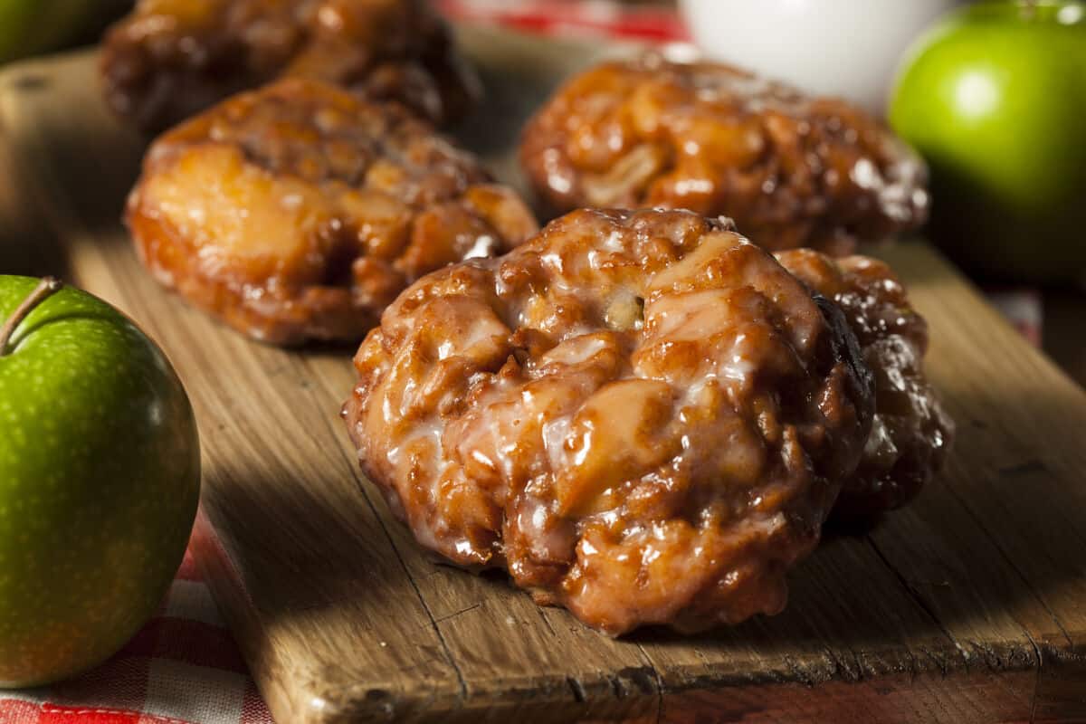 3 large apple fritters on a wooden cutting board, with some whole green apples on the side, almost out of s.