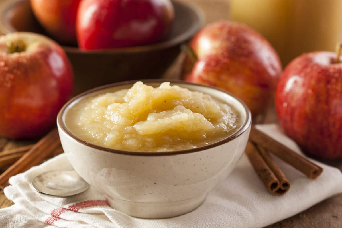 A white bowl full of thick apple sauce, in front of some apples in a blurred backgro.