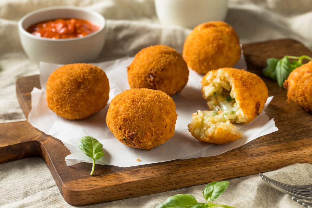 5 arancini on a wooden cutting board, with one split open to show the inside. A bowl of tomato based dip can be seen in the blurred backgro.