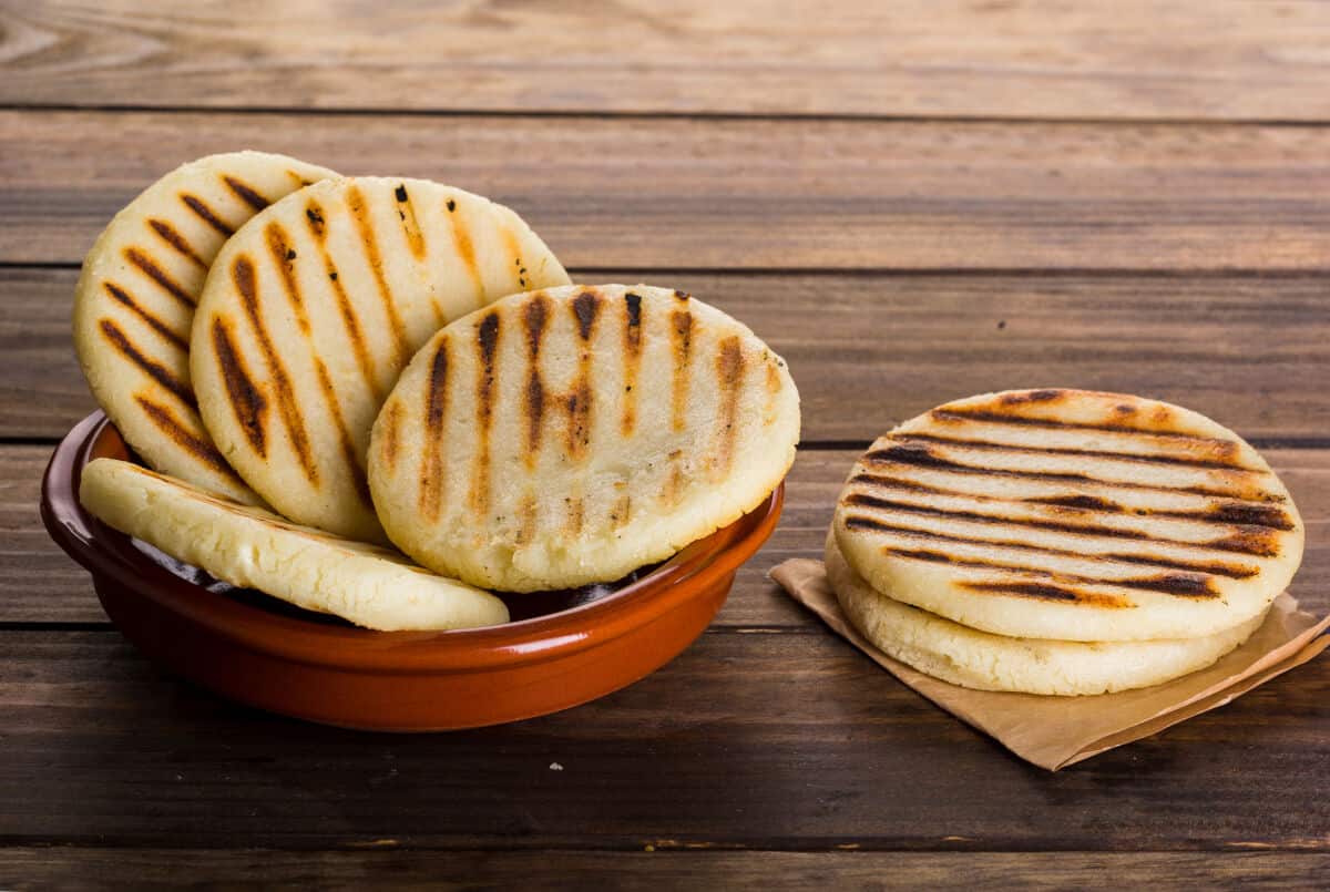 A bowl full of 4 arepas, next to 2 more on a folded napkin on a wooden ta.