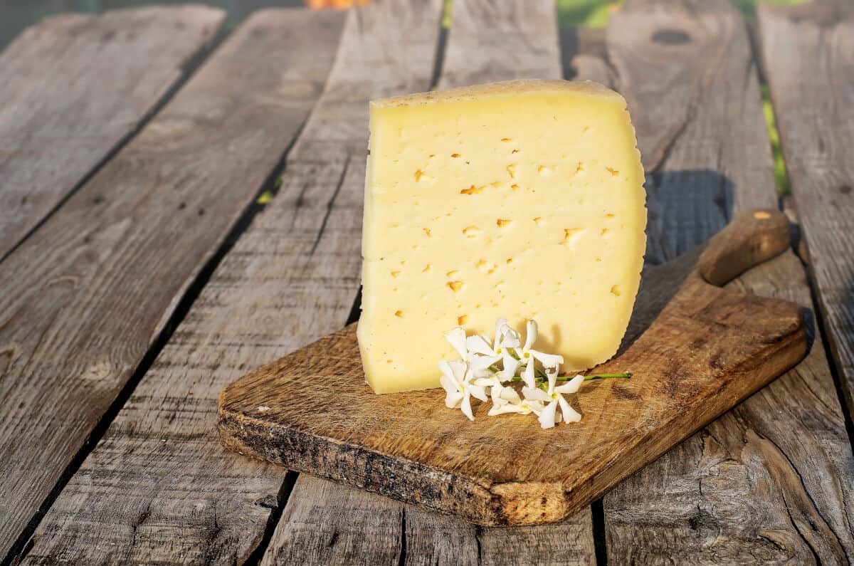 A triangle of asiago cheese on a wooden paddle cutting board, with some tiny white flowers in fr.