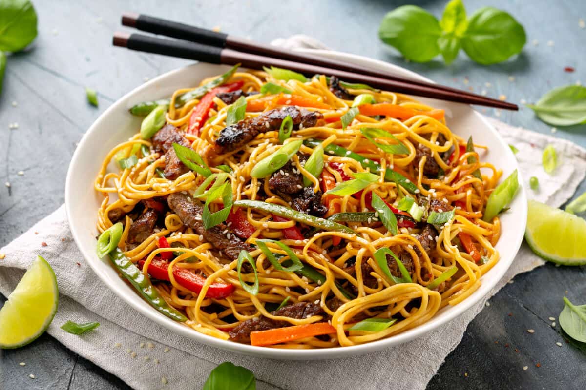 A bowl of Asian noodles, with chopsticks resting on the lip of the bowl, and some lime quarters scattered aro.