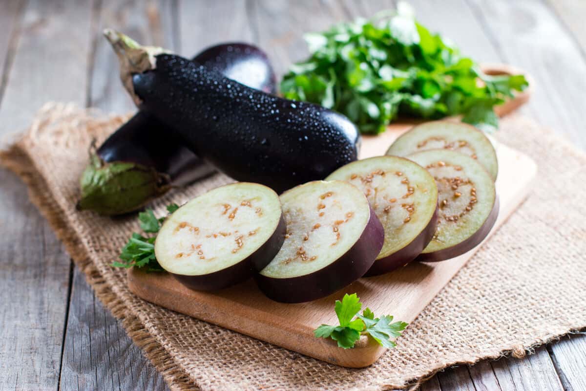 5 aubergine slices fanned out in front of two whole aubergine, on a wooden paddle board, on a hessian cl.