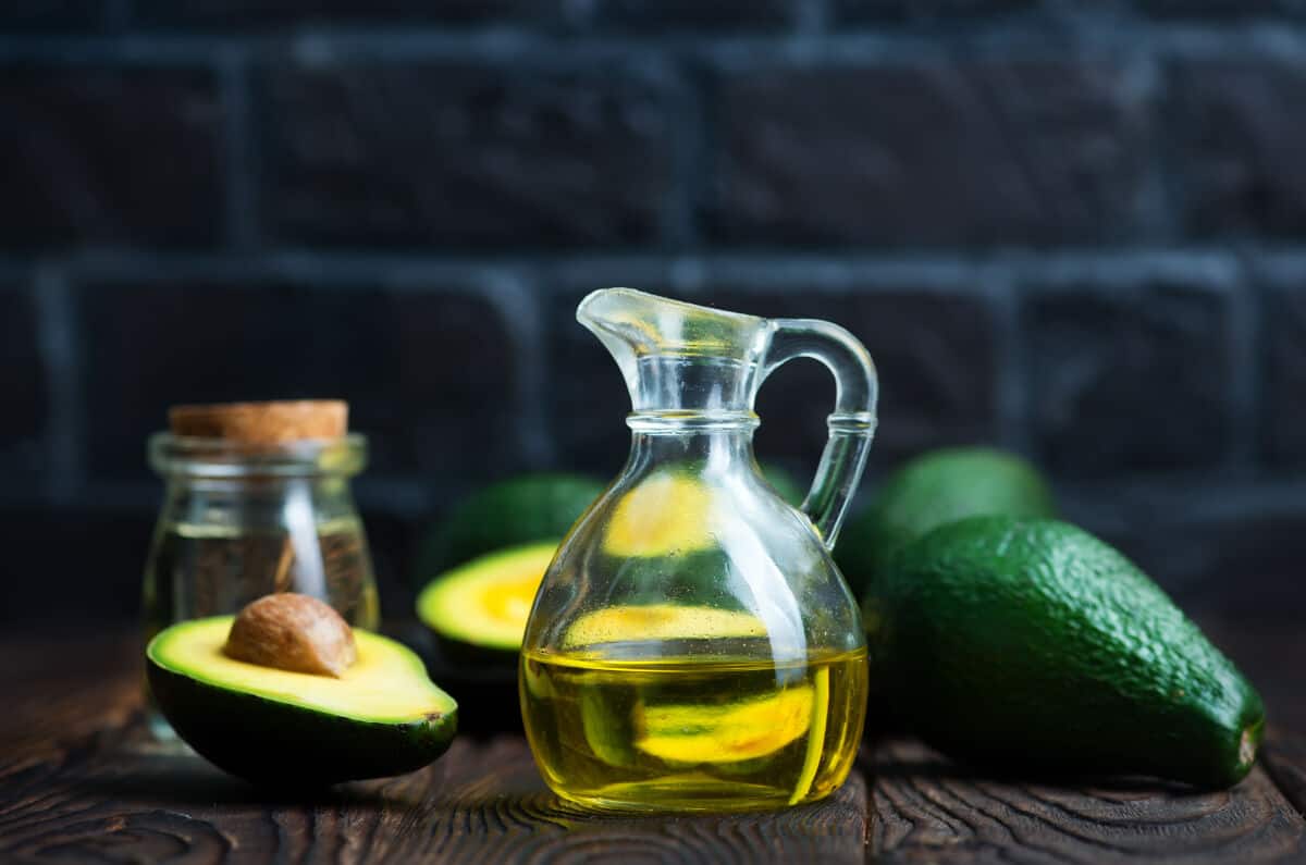 A small glass jug half full of avocado oil, in front of some whole and some half avoca.