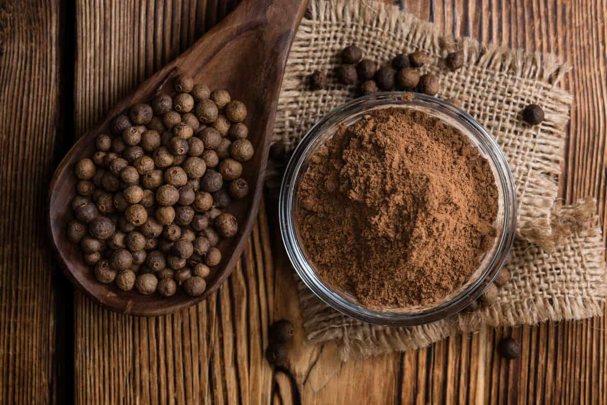 A bowl full of whole allspice, next to a wooden scoop full of powdered allspice, on a piece of hessian on a dark wooden ta.