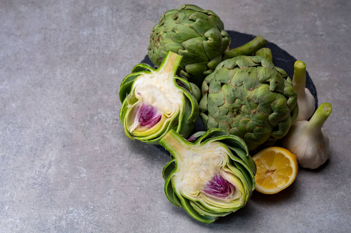 Two whole and one halved artichokes on a greyish stone looking surf.