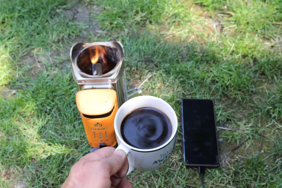 A Biolite CampStove with flames coming out the top, a cup of coffee in a mans hand, and a charging mobile phone, all on grass.